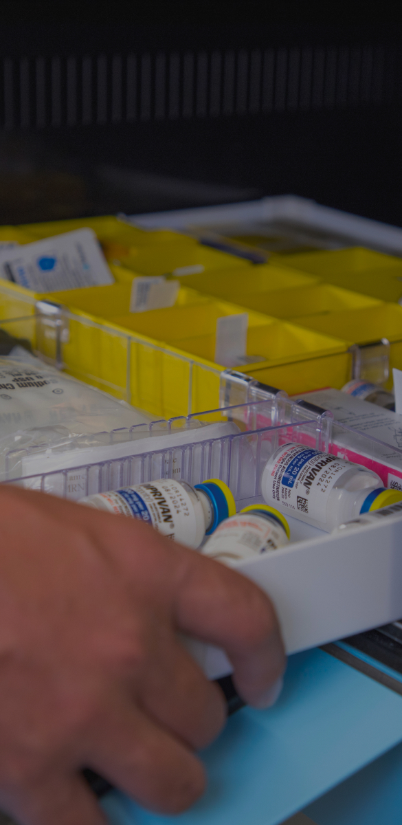 A hand reaches for a tray of vials in a storage unit, with yellow compartments and various medical supplies visible.